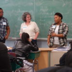 Three speakers stand against a chalkboard talking to students. "LGBT" is visible on the chalkboard.