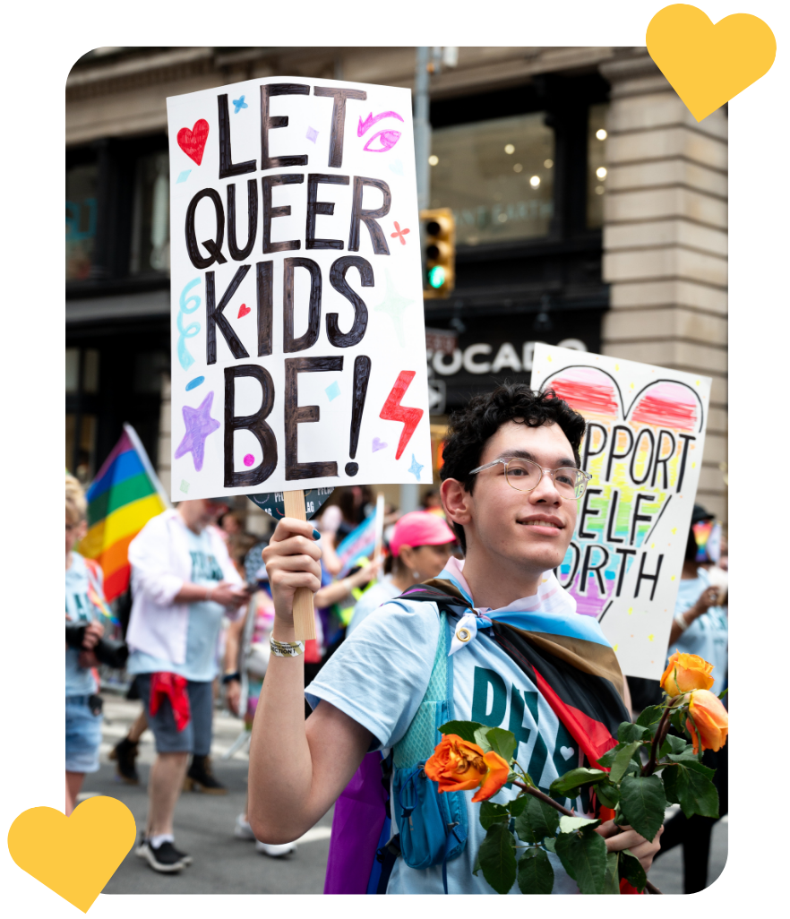 An LGBTQ+ youth holds roses in one hand and a homemade sign that reads, "Let queer kids be!" in the other. They are wearing a light blue PFLAG NYC tshirt while wrapped in a progress Pride flag.