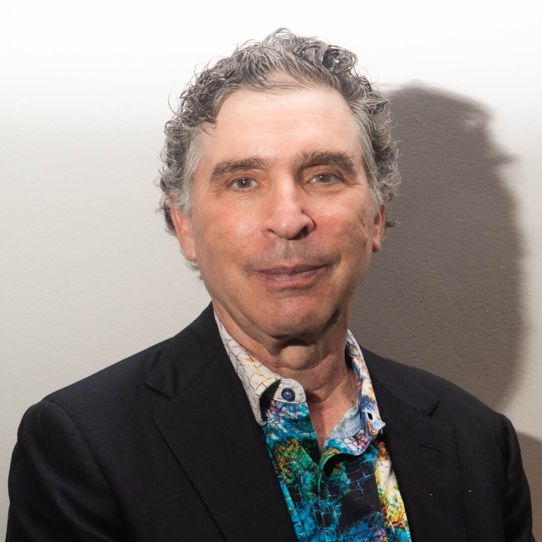 A headshot of PFLAG NYC Board Member, Lee Arthurs, who wears a dark suit jacket and a multicolored collared shirt beneath it