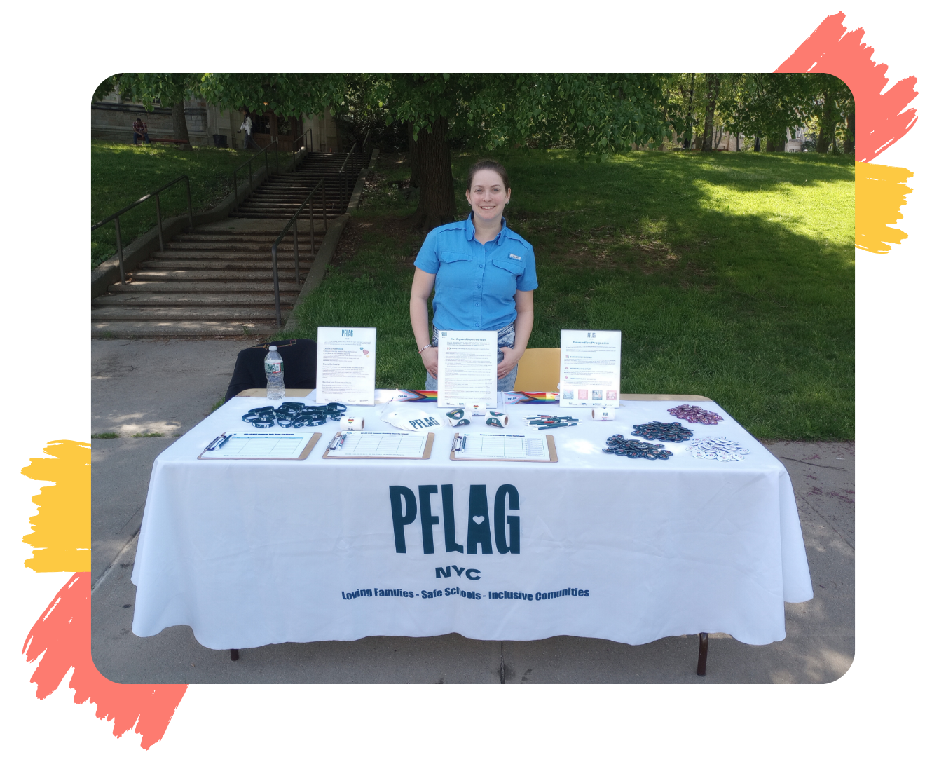 A staff member named Shelby stands behind a table covered with a white tablecloth displaying the PFLAG NYC logo. The table is set up outdoors in a park with green grass and trees in the background. On the table are various informational materials and promotional items such as wristbands and buttons. The surrounding border of the image has artistic brush strokes in red and yellow on the corners.