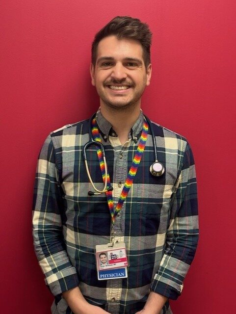 A headshot of Dr. Birkhead in front of a red background wearing a rainbow striped lanyard.